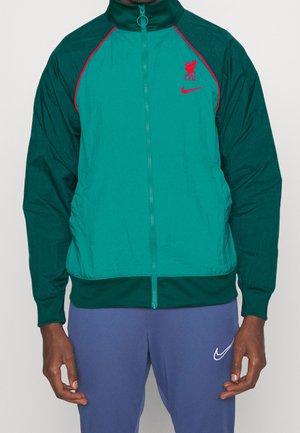 Homme portant une veste zippée Liverpool FC turquoise et vert foncé avec un logo rouge, et un pantalon Nike bleu avec un logo blanc.