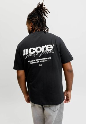 Mand med dreadlocks iført en sort t-shirt med hvid tekst, der lyder "JJCORÉ Athletic Club Worldwide Global Movement Co.", og grå bukser.