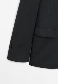 Mörk rutmönstrad kavajärm med en slät textur, utrustad med två svarta knappar och en kavajslag med skåra. Skräddarsydd passform med en strukturerad design.