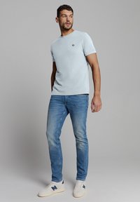 Hellblaues T-Shirt mit kurzen Ärmeln und rundem Ausschnitt, versehen mit einem kleinen Logo, kombiniert mit mittelblauen Jeans und weißen Sneakers.