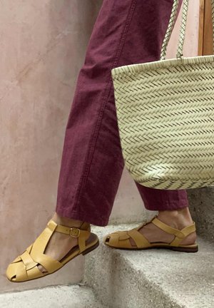 Osoba koja nosi kožne sandale u boji goruće žute i bordo hlače penjući se kamenim stepenicama, držeći pletenu torbu od slame.