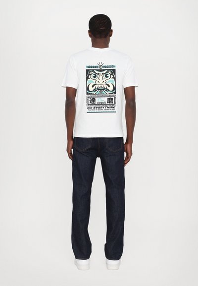 Camiseta de algodón blanca con un gran diseño gráfico de una cara en azul, negro y blanco. Combinada con jeans de mezclilla oscura y zapatillas blancas.