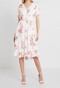 Blommig midiklänning med v-ringning, korta ärmar, vit färg och midjeband. Har rosa och orange blommor mot en ljus bakgrund.