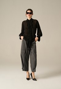 Blusa nera trasparente con dettaglio frontale a ruches, abbinata a pantaloni grigi a gamba larga in stile cargo. La modella indossa occhiali da sole neri oversize e tacchi.