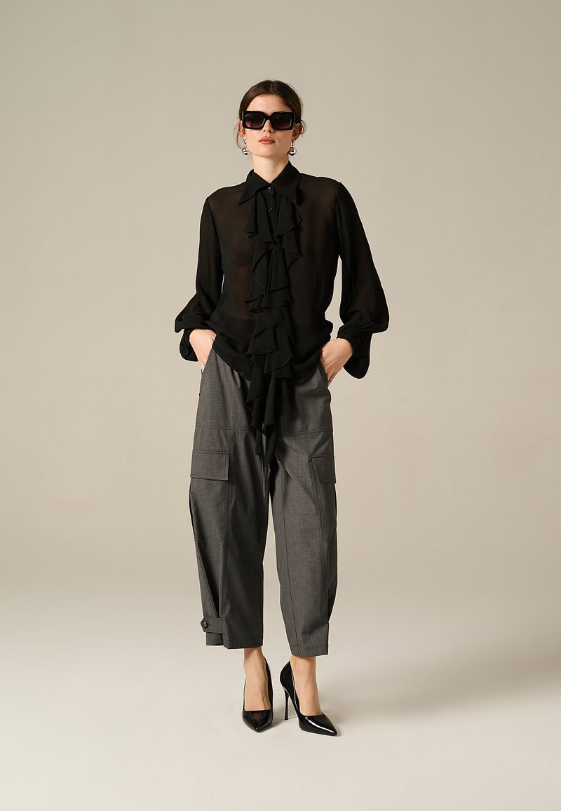 Blusa nera trasparente con dettaglio frontale a ruches, abbinata a pantaloni grigi a gamba larga in stile cargo. La modella indossa occhiali da sole neri oversize e tacchi.