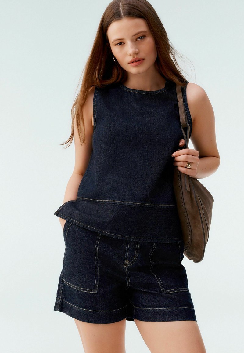 Tmavě modrý bezrukávový top s peplum lemem, přičemž je kombinován s odpovídajícími kraťasy. Jednoduchý design s viditelným prošíváním a uvolněným střihem.