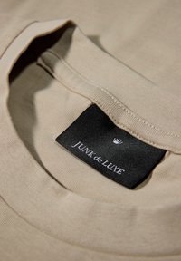 Beige bomulds t-shirt med flad krave; har et sort mærke med teksten "JUNK de LUXE" og et krone-logo ovenfor. Glat tekstur.