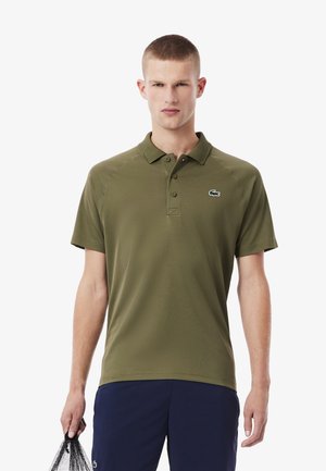 Camisa polo verde oliva con un cuello clásico, una tapeta de tres botones y un pequeño logotipo bordado en el lado izquierdo del pecho. Textura de tela suave.