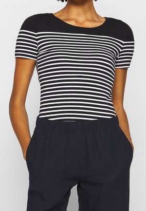 Persoon die een zwart-wit horizontaal gestreept shirt met korte mouwen en een zwarte broek draagt, met handen in de zakken.