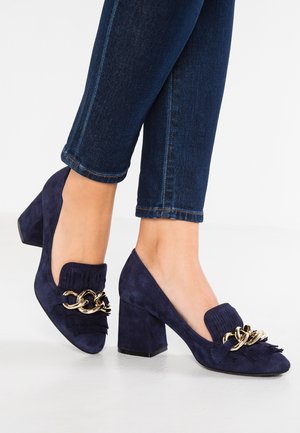 Navy blauwe suède loafers met blokhak en gouden kettingdetail, gedragen met donkerblauwe denim jeans, getoond vanaf halverwege het kuitbeen naar beneden.