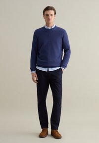 Navy blue sweater, textured knit, crew neck, layered over a light blue button-down shirt, paired with dark pants and brown shoes.