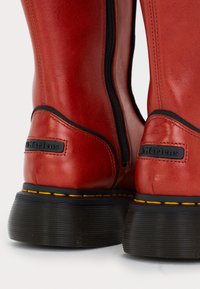 Rode leren laarzen met zwarte dikke zolen, gele stiksels en zwarte ritslijnen, met het logo-tab aan de achterkant van de hak.