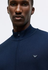 Pulover cu fermoar navy, realizat dintr-un material tricotat texturat. Are un guler înalt și un mic logo pe piept.