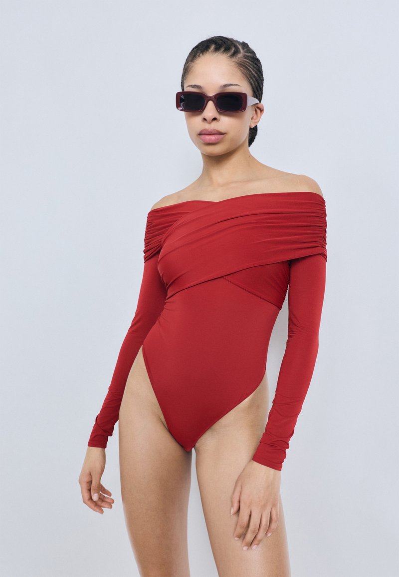 Punane üheõlaline ühtne ujumisriietus, millel on pikad varrukad, volditud disain ja kõrge lõikega jalaava. Sile, elastne kangas.