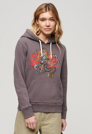 Frau trägt einen grauen Hoodie mit buntem Drachenmuster und beigen Hosen, steht vor einem schlichten hellen Hintergrund.