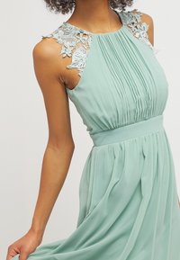 Vihreä pleatattu mekko, jossa on pitsiyksityiskohtia olkapäillä ja vyötärö on istuva. Kankaassa on virtaava laatu ja pehmeä rakenne, sekä elegantti siluetti.