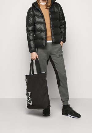 Veste matelassée noire portée sur un pull marron, pantalon de survêtement gris avec accents noirs, et un sac fourre-tout noir avec le logo "EA7", assortis à des baskets noires.