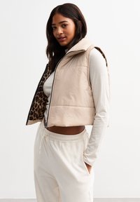 Beige puffer vest with high collar, front zip and animal print lining, worn over a light-colored long-sleeve shirt and cream joggers.