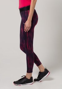 Dopasowane legginsy w ciemnofioletowym materiale z czerwonymi pionowymi paskami. Posiadają szeroki czarny pas w talii i są zestawione z czarnymi butami sportowymi.