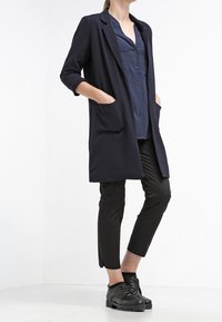 Marineblå lang blazer med åben front, enkelt knaplukning og to forlommer, parret med sorte cropped bukser og teksturerede sorte sko.