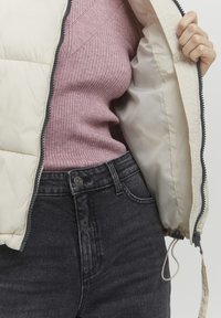 Lätt pufferjacka i creamfärg, ozipprad, visar en ribbad rosa tröja under, tillsammans med mörk denimjeans med en knapp framtill.