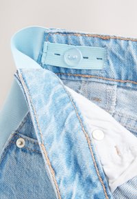 Lyseblå denimjeans med en blød elastisk linning, knapdetalje og kontrastsyning nær lommer og lynlåsområde.