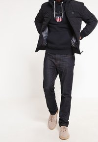 Schwarze gesteppte Jacke, marineblauer Kapuzenpullover mit Logo, dunkle Jeans, beige Schuhe. Der Kapuzenpullover hat eine Vorderentasche; die Jacke ist mit grauem Futter ausgestattet.