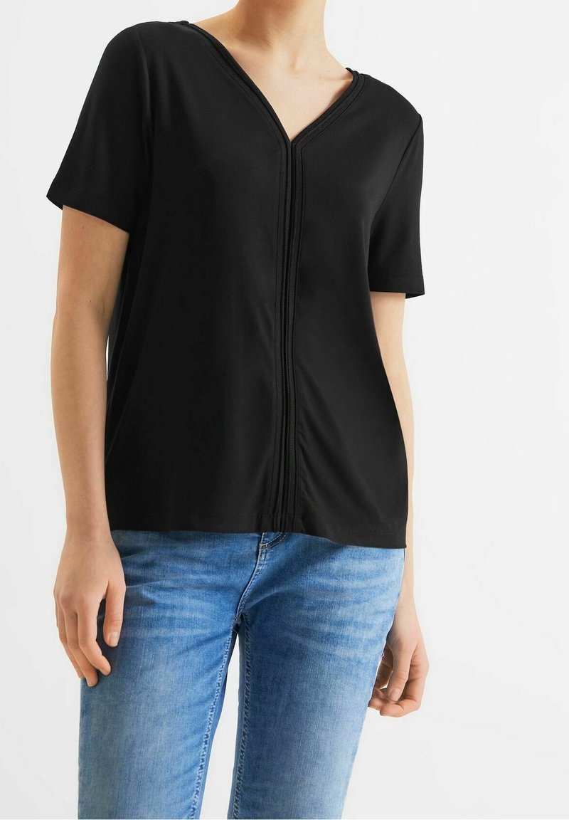 Top noir à manches courtes avec un col en V et un détail de couture texturé descendant sur le devant. Assorti à un jean bleu, mettant en valeur une coupe décontractée.