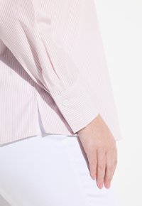 Camicia a righe rosa chiaro con polsini abbottonati, caratterizzata da maniche lunghe e una vestibilità rilassata. Indossata su pantaloni bianchi, evidenziando il contrasto.