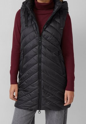 Nainen, joka käyttää mustaa tikattua hihatonta liiviä, jossa on etuvetoketju, päällään viininpunainen poolopaita ja harmaat housut, seisoo yksiväristä taustaa vasten.