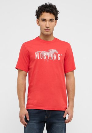 Junger Mann mit lockigem Haar, der ein rotes Mustang-T-Shirt und dunkelblaue Jeans trägt, vor einem einfarbigen hellen Hintergrund stehend.