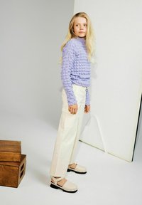 Haut à manches longues texturé violet, pantalon cargo crème et sabots beiges avec des semelles noires. Le mannequin se tient avec un bloc en bois en arrière-plan.