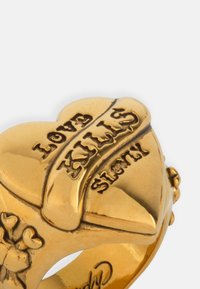 Heart-shaped gold ring with engraved "Love Kills Slowly" design and small heart accents on the sides. Smooth, shiny surface with rounded edges.
