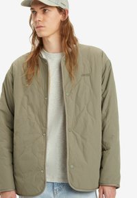 Quilted olive green jacket featuring snap buttons, ribbed trim, and an embroidered logo on the chest, worn over a gray shirt.