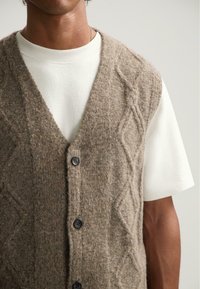 Mann, der eine beigefarbene strukturierte Strickweste mit Knöpfen über einem schlichten weißen Kurzarmhemd trägt, vor neutralem Hintergrund.
