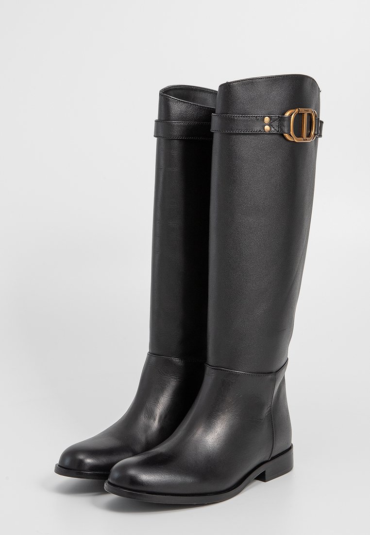 Stivali in pelle neri al ginocchio con punta arrotondata e cinturini con fibbie dorate vicino alla parte superiore, presentati su uno sfondo chiaro e semplice.