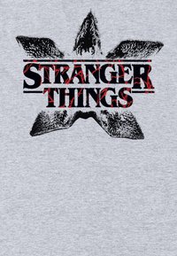 Szara koszulka z przetartym czarno-czerwonym logo "Stranger Things", otoczonym symetrycznym wzorem graficznym przypominającym skrzydła nietoperza.