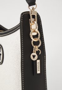 Zbliżenie rogu czarno-białej torebki Guess z złotym łańcuchem i detalem z metką na białym tle.