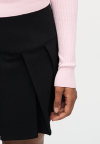 Main droite reposant près de l'ourlet d'une jupe noire, avec un pull à manches longues côtelé rose clair visible sur le bras.