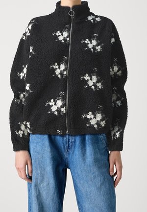 Schwarze Fleecejacke mit Reißverschluss und weißer Blumenstickerei, getragen mit blauen Jeans, mit langen Ärmeln und hohem Kragen.