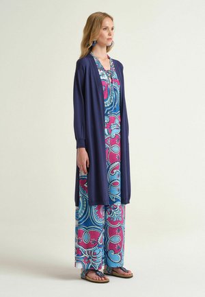Donna che indossa un lungo cardigan blu navy sopra pantaloni a gamba larga con motivi blu e rosa e un top coordinato, abbinati a orecchini blu e sandali.