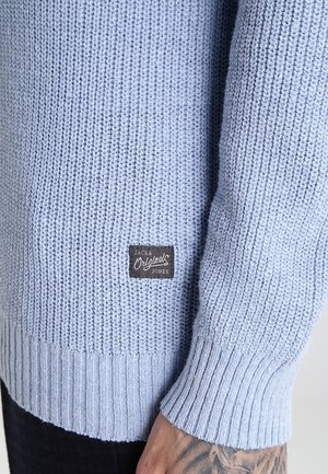 Manche et ourlet d'un pull tricoté bleu clair avec une étiquette noire "Jack & Jones Originals" au-dessus d'un poignet et d'une main tatoués.