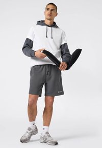 Felpa grigia con accenti bianchi e grigi scuri, abbinata a short grigi. Il modello tiene due accessori neri. Lo sfondo è bianco.