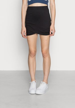 Kvinde iført en sort omslagsmini nederdel, hvide platform sneakers og en blå cropped top, der står mod en ensfarvet baggrund.