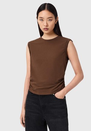 Młoda kobieta o prostych ciemnych włosach ubrana w brązowy bezrękawnik i czarne jeansy, stojąca z jedną ręką w kieszeni na szarym tle.