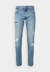 Jean en denim bleu clair avec des patchs usés sur la cuisse gauche, le genou droit et au-dessus de la cheville droite, présenté sur un fond blanc.