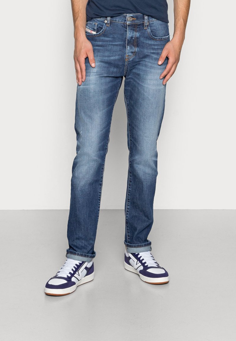Blue denim jeans with a straight-leg design, faded finish, and classic five-pocket style. Paired with white and navy sneakers.