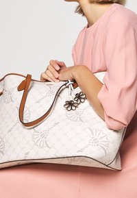 Vit handväska med ett tonalt mönster, bruna läderhandtag och blommig charmdetalj. Väskan har en strukturerad form med en slät yta.
