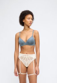 Γυναίκα με σγουρά μαλλιά που φοράει μπλε σουτιέν και ζώνη για καλτσοδέτες από κρεμ δαντέλα με λουριά, σε απλό ανοιχτό φόντο.