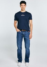 Jonge man met één hand in de zak, gekleed in een marineblauw "Authentic" T-shirt, blauwe spijkerbroek, bruine riem en witte sneakers tegen een effen achtergrond.